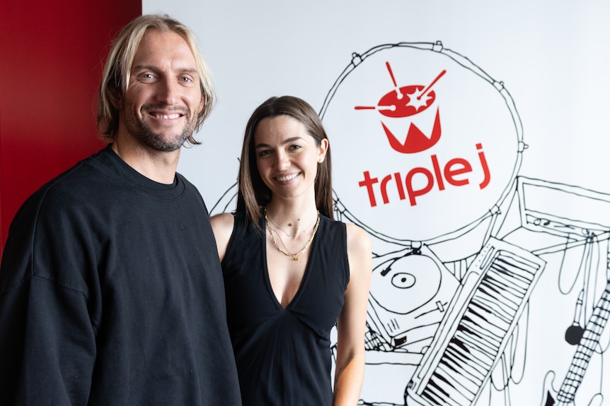 a man and a woman smile at the camera standing in front of a triple j sign 