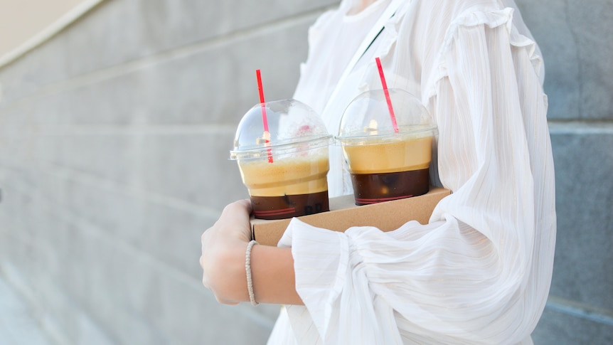 A woman wearing a white blouse holds a cardboard carry tray containing two takeaway ice lattes in plastic cups with domed lids.