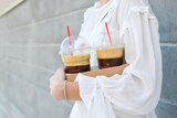 A woman wearing a white blouse holds a cardboard carry tray containing two takeaway ice lattes in plastic cups with domed lids.