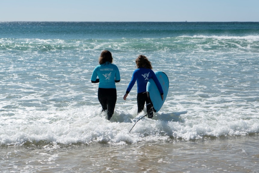 A woman and a child walk into the surf with a surfboard for a story about surf therapy for autistic children