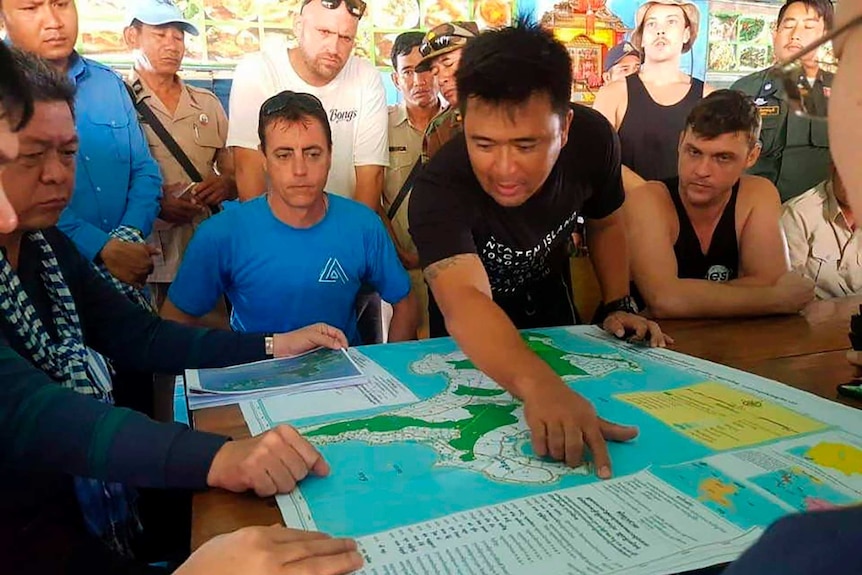 A man surrounded by a group of people points at a map on a table.