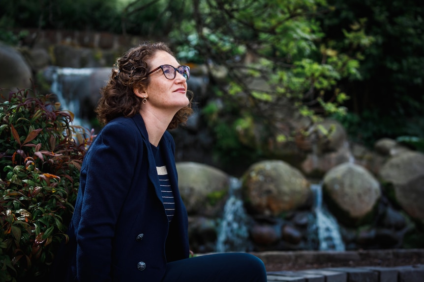 A woman with short curly hair, wearing a navy blazer, sits by a small water feature, looking up to the trees