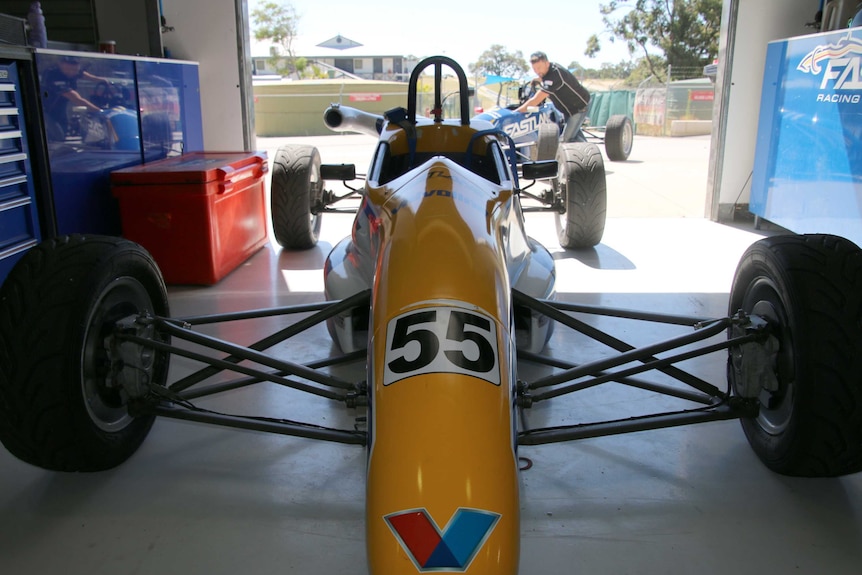 A yellow racing car sits in a garage while a mechanic wheels a blue car in the background.