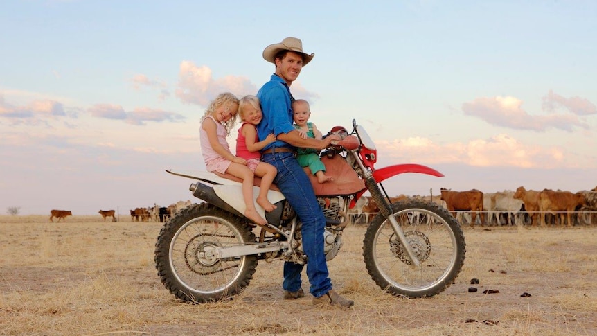 A man wearing a cowboy hat, sitting on a bike with three children with cows in the background