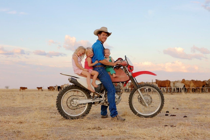 A man wearing a cowboy hat, sitting on a bike with three children with cows in the background