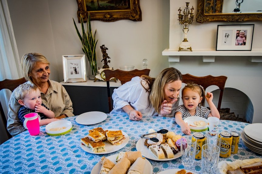 Mathew sits on Rose's lap at the dinner table, while Robyn speaks to Emilia who is reaching for some food.