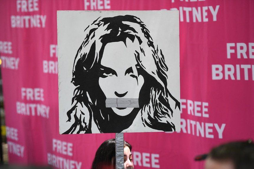 A black-and-white poster of Britney Spears is held in front of pink backdrop reading FREE BRITNEY