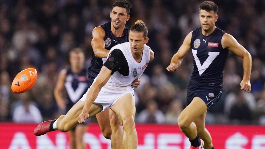 two men in Victoria jumpers behind a man in a white jumper with an orange AFL ball in front of him