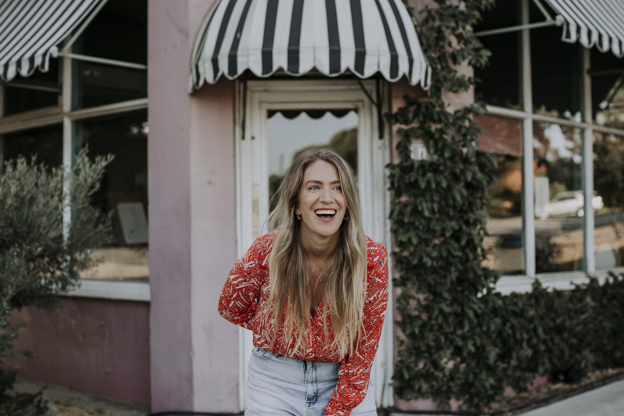 A woman bending forward with laughter outside a pink building with vines creeping up the wall.