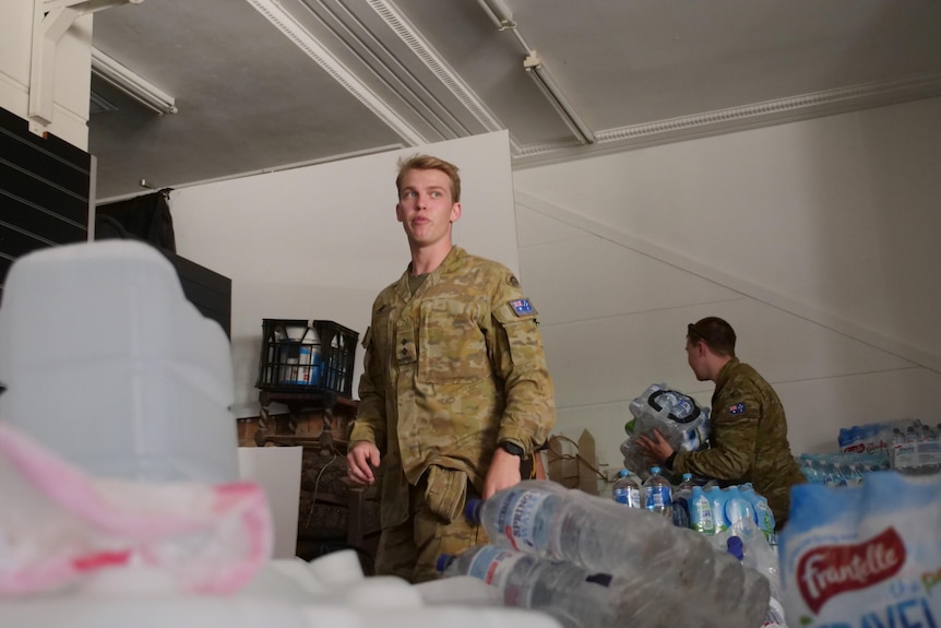 A man in a army uniform stands in a room full of water bottles while another man picks up a pack of bottles.