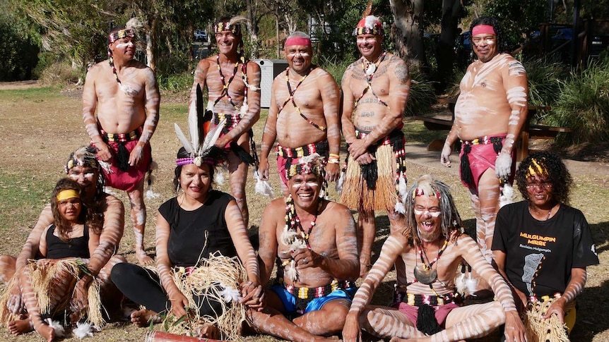 A group of men and women dressed in traditional indigenous clothes and paint stand in a park and smile at the camera.