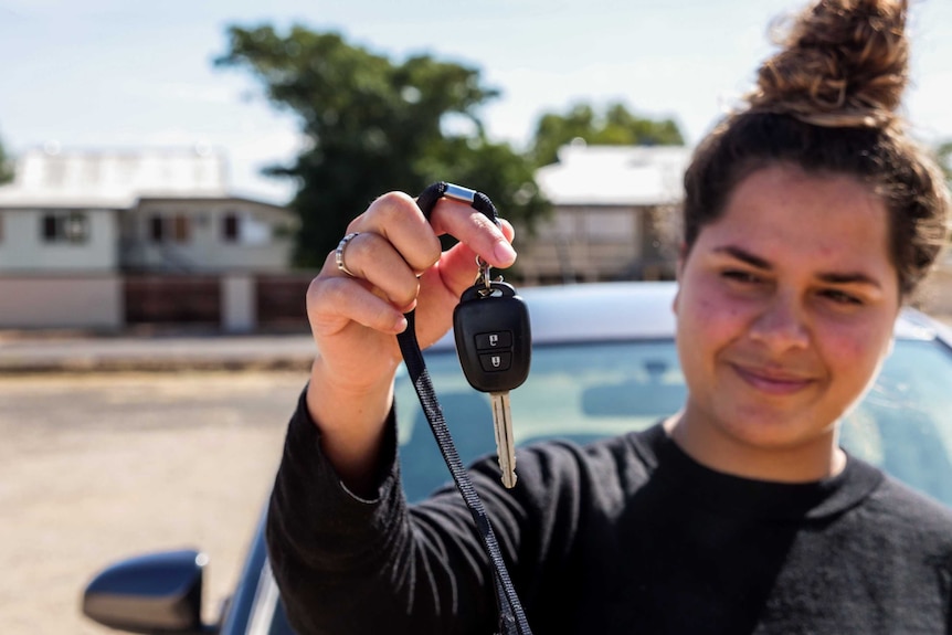 A teenager holds out a car key, the key is in focus and she is not. Her hair is in a bun.