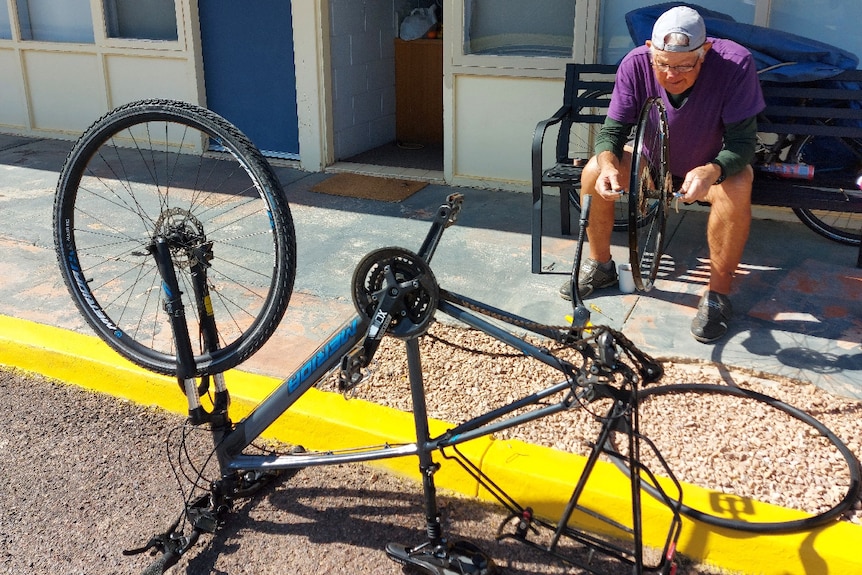 Del sitting on chair outside motel room examining bike wheel in his hands, bike upside down in foreground