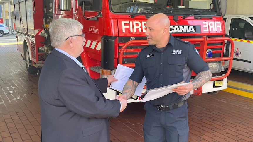 Two men, one in a suit and one in a firefighter uniform, shake hands and pass a sheet of paper.