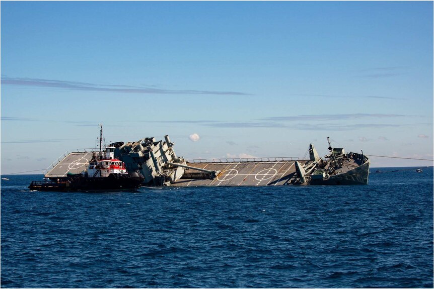 A navy ship tilts to one side on the ocean, with a tug boat on its side.