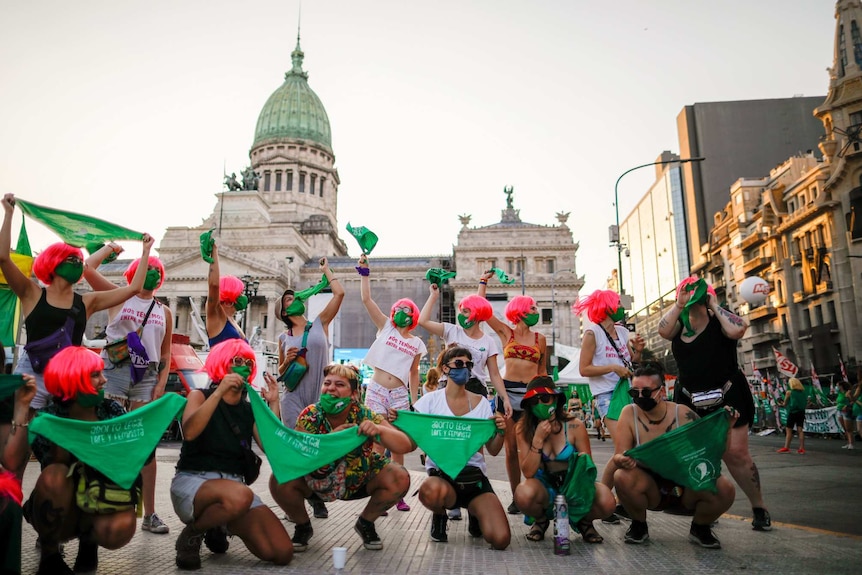 A group of women wearing pink wigs, green masks and holding green flags stand or squat outside a building.