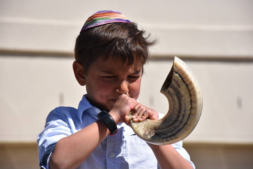 A small boy wearing a blue shirt and yamulke blows a horn.