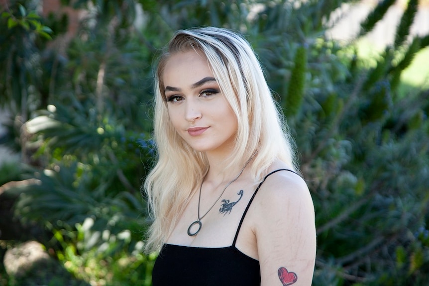 A woman stands in front of some greenery dressed in a black dress