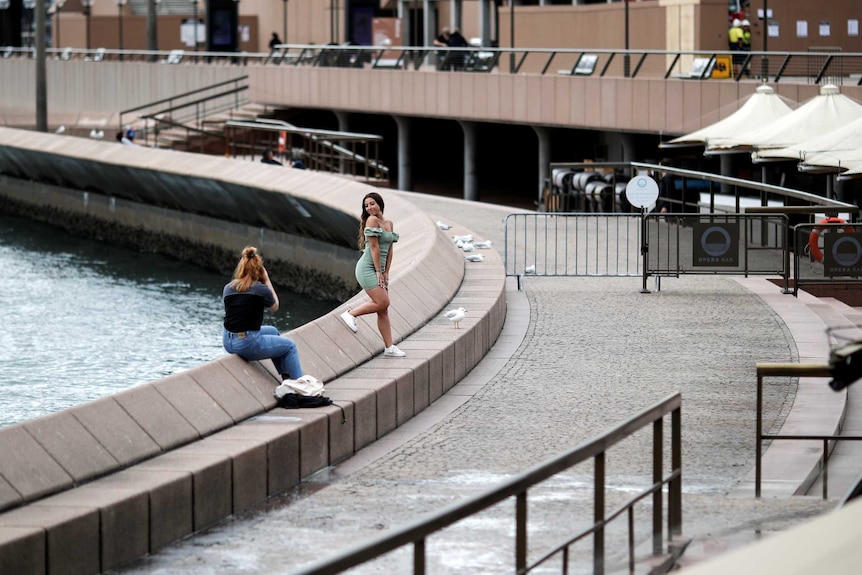 A woman takes a photo of another woman posing outside the Sydney Opera House which is deserted