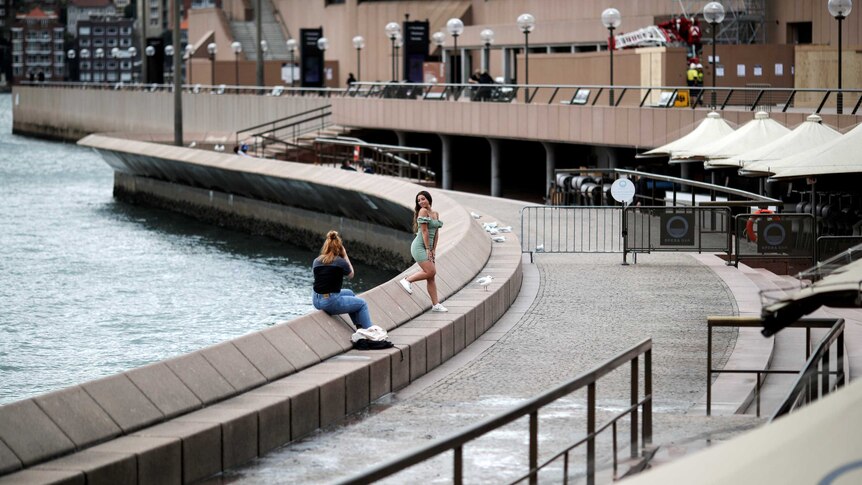 A woman takes a photo of another woman posing outside the Sydney Opera House which is deserted