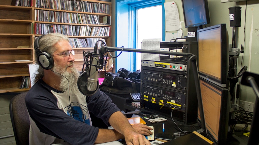 A man with grey hair and a long beard sits behind a radio studio desk, headphones on.