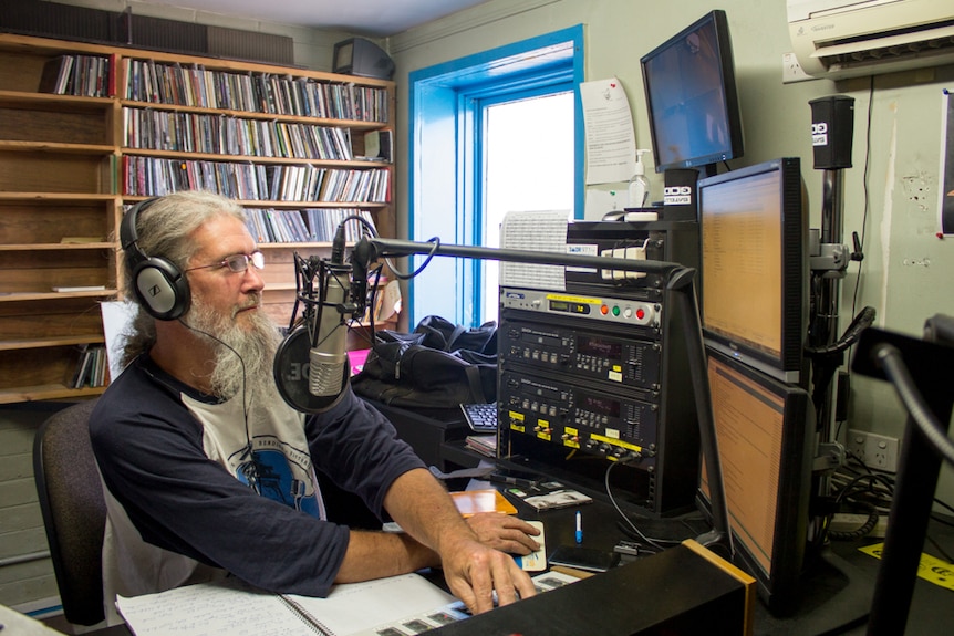 A man with grey hair and a long beard sits behind a radio studio desk, headphones on.