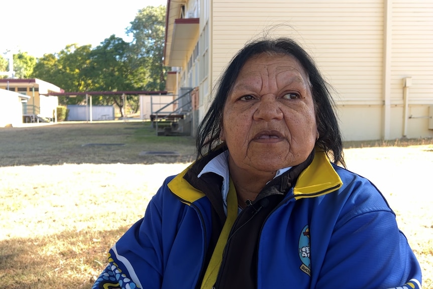 An older Indigenous woman in yellow and blue jacket stands in front of sun-lit buildings, looks away from camera.