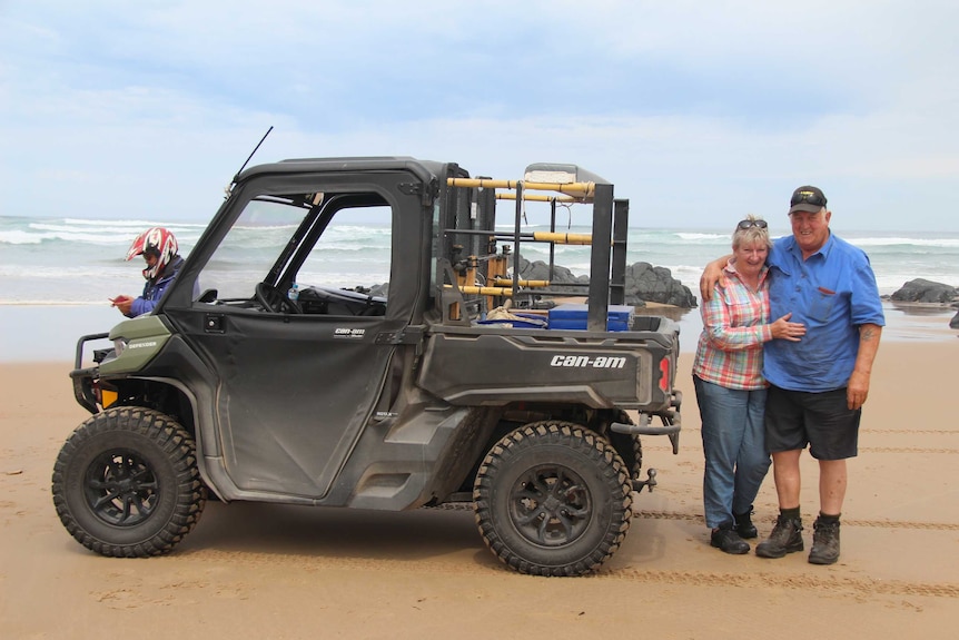 A couple in their sixties with arms around in other standing near rugged beach mobile on wide beach with surf behind