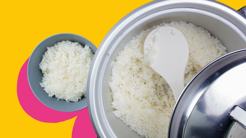 A rice cooker is seen with rice in it with the lid in the middle of being taken off. A spoon sits inside, a bowl is on the left.