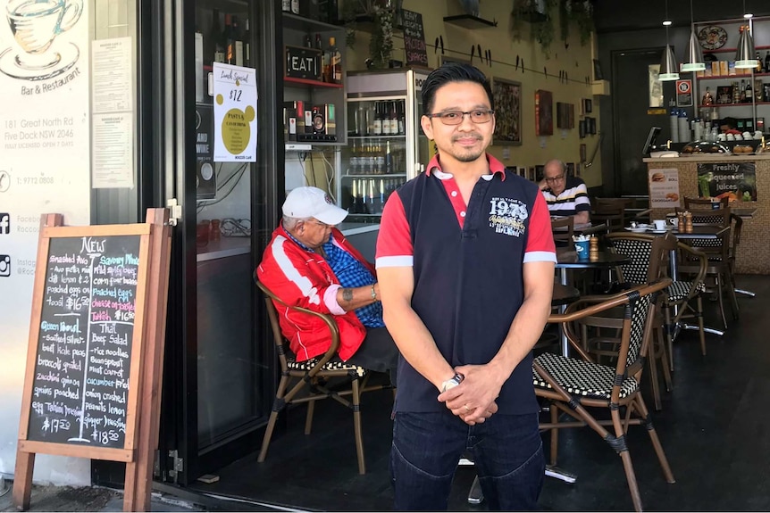 A man wearing an apron stands outside a cafe.