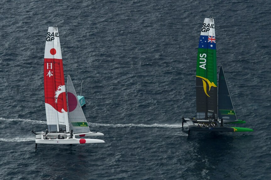 A sailing boat with green sails with a gold kangaroo motif sails away from another boat, with white and red sails