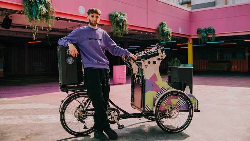 A man in purple jumper stands beside a customised bicycle with DJ decks built in