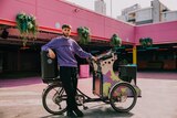 A man in purple jumper stands beside a customised bicycle with DJ decks built in