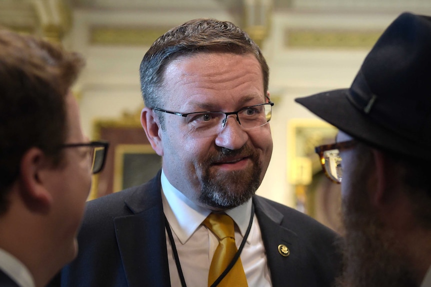 A close-up of Sebastian Gorka wearing a suit smilies while talking to two men either side of him