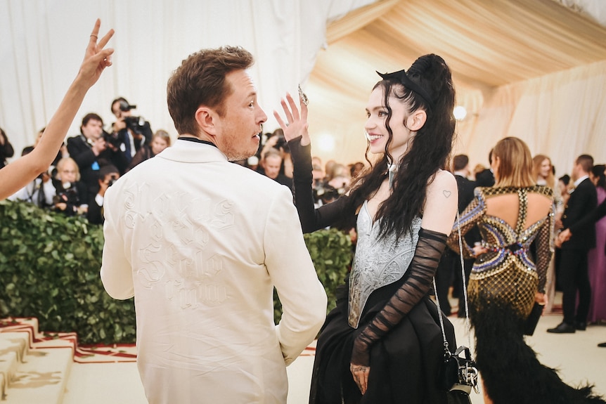 Elon Musk vistiendo un traje blanco con su pareja Grimes, quien viste mayormente de negro