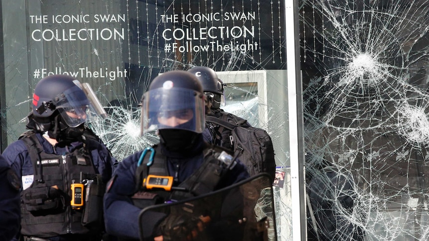Three riot police in black helmets stand in front of a large glass window that is smashed.