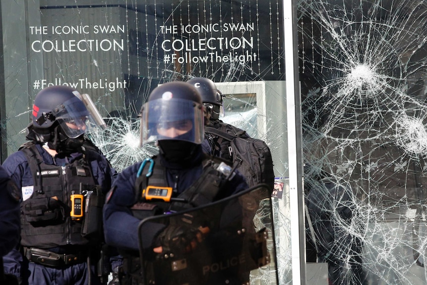 Three riot police in black helmets stand in front of a large glass window that is smashed.