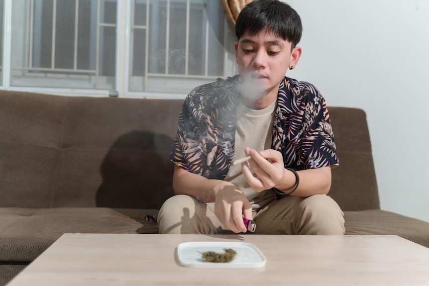 A young man sits on the couch smoking a joint in front of an ashtray