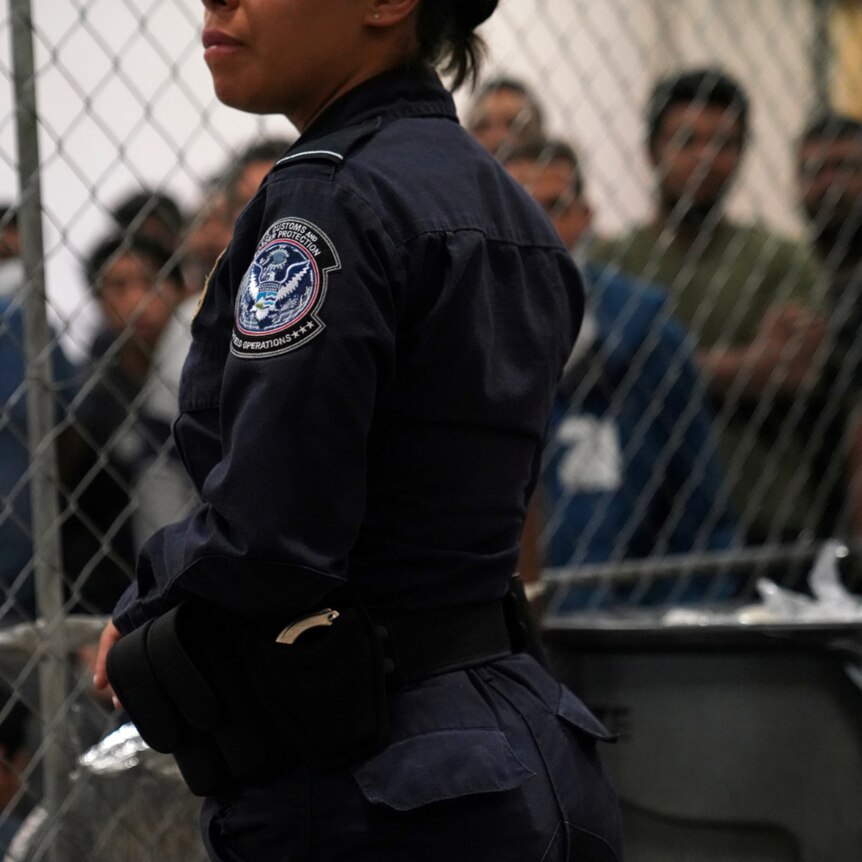 A woman in a navy Border Control uniform stands before a chain link fence with men behind it.