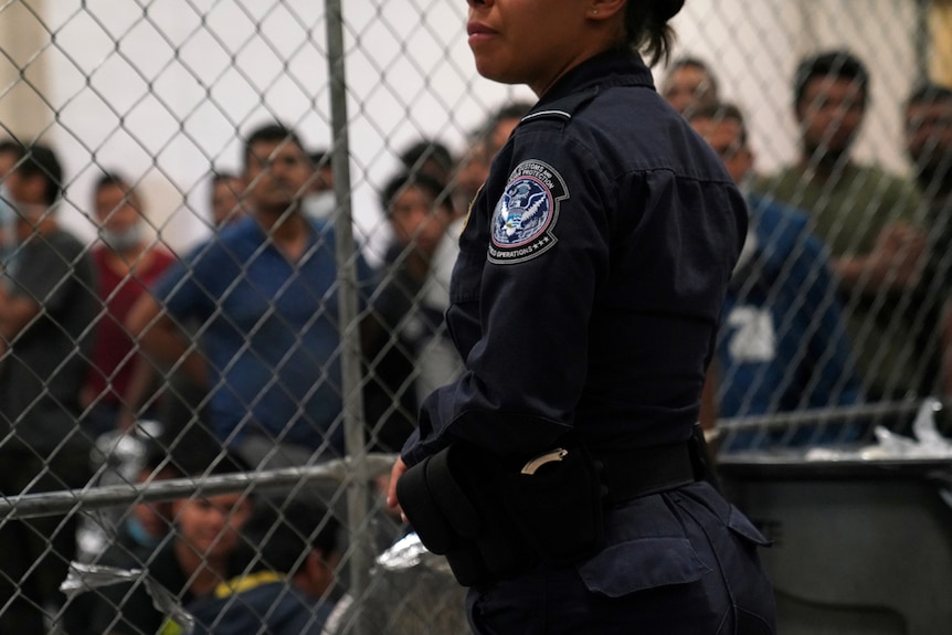 A woman in a navy Border Control uniform stands before a chain link fence with men behind it.