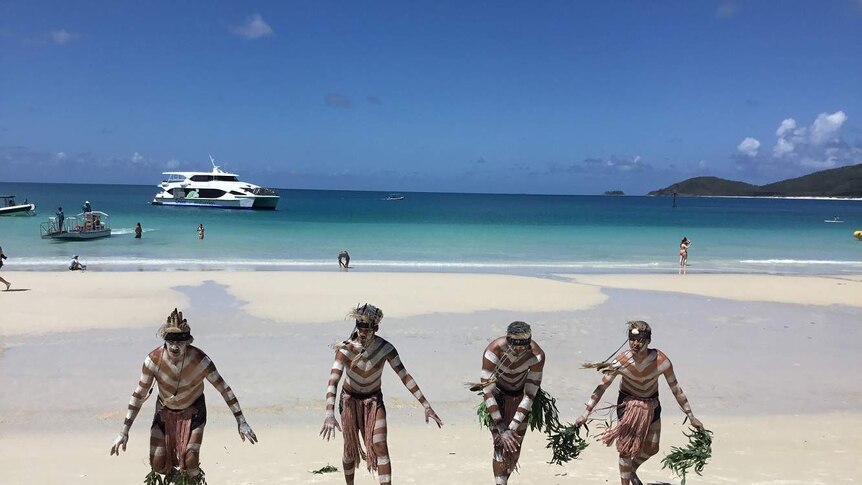 Group of Indigenous man in traditional dress dance on a white sandy beach.