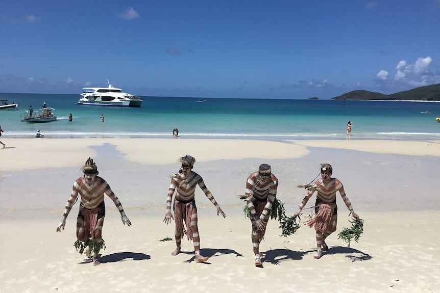 Group of Indigenous man in traditional dress dance on a white sandy beach.