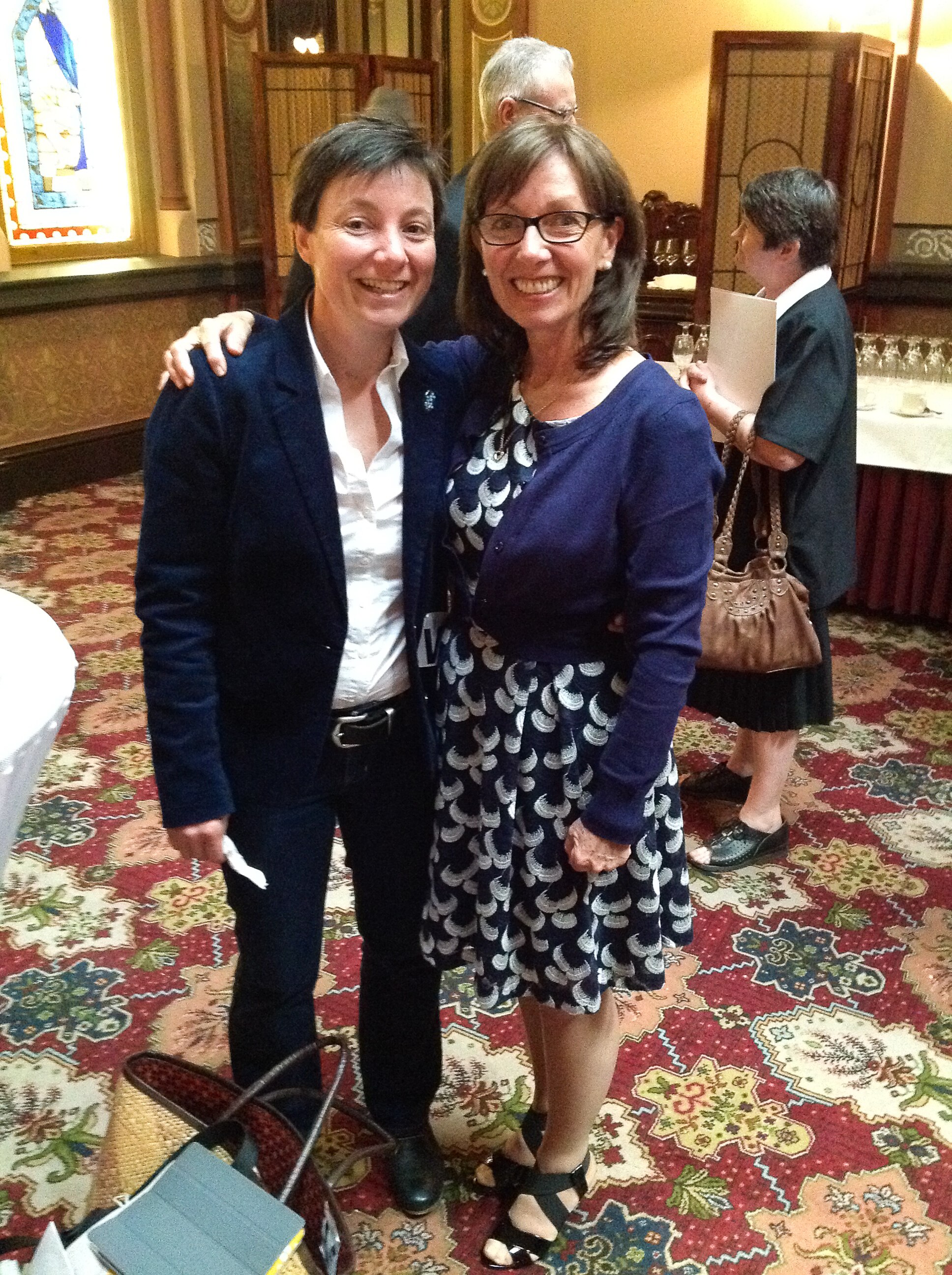 Two women stand in a parliamentary lobby wearing semi-formal attire, smiling. 