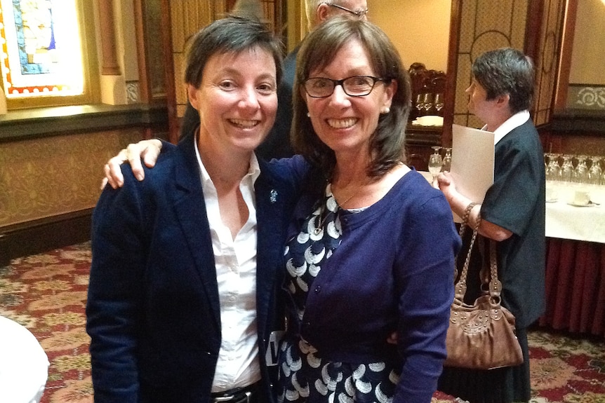 Two women stand in a parliamentary lobby wearing semi-formal attire, smiling. 