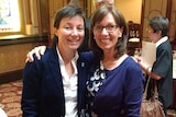 Two women stand in a parliamentary lobby wearing semi-formal attire, smiling. 