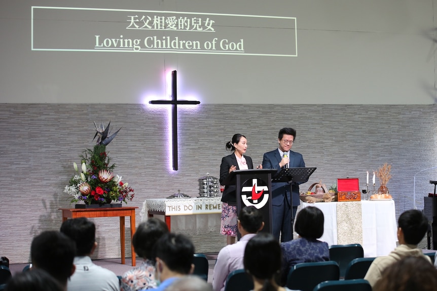 A woman and man stand at a pulpit in front of a church congregation