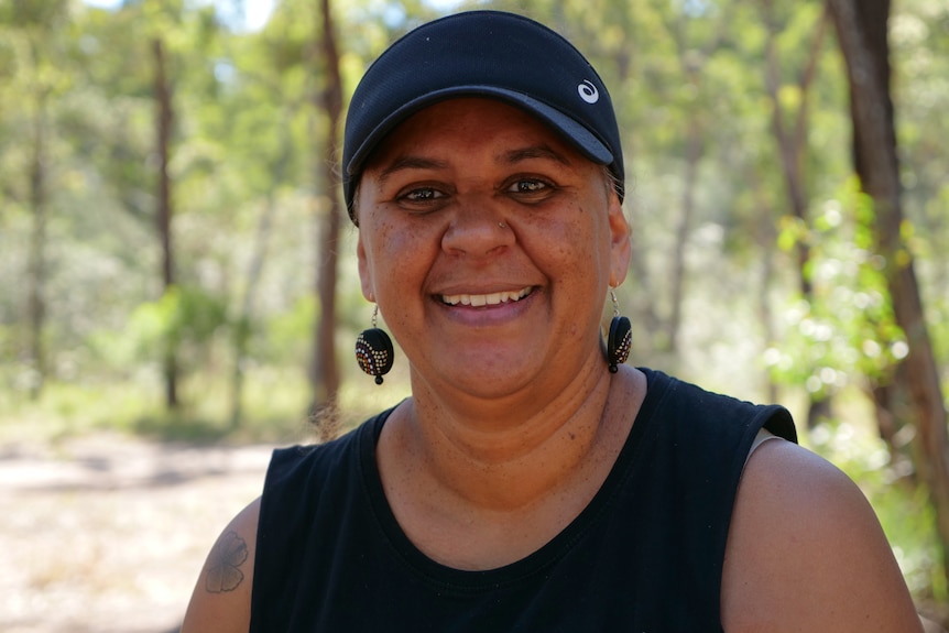 Esther Lampton, smiling, colourful earrings, black cap, trees behind.