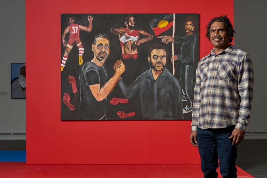 Artist Vincent Namatjira standing smiling next to his Archibald Prize winnining painting of footballer Adam Goodes