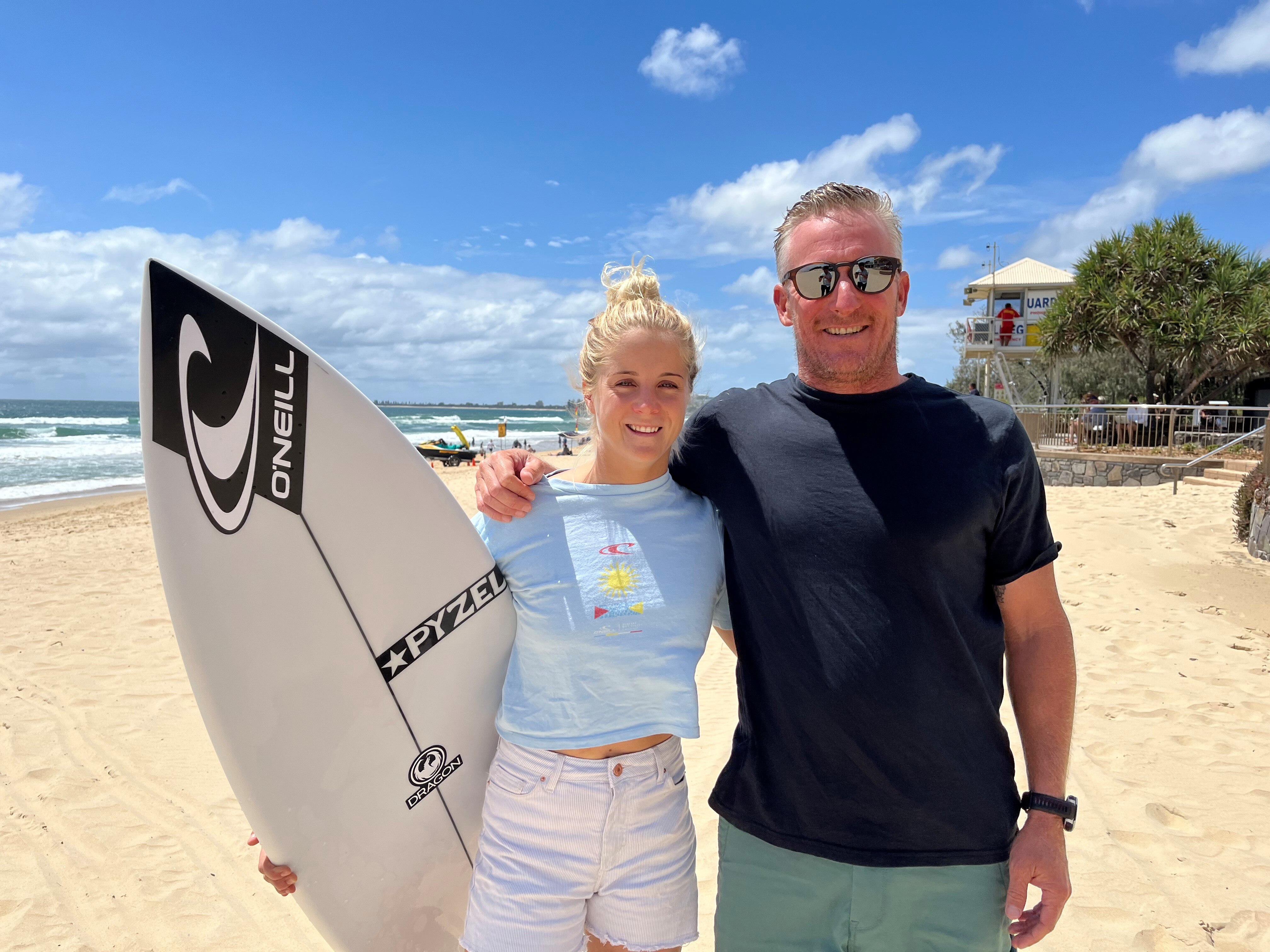 Woman on beach holding surfboard with male coach