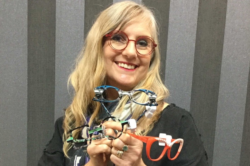 A woman wearing red framed glasses smiling while holding three pairs of bright coloured glasses